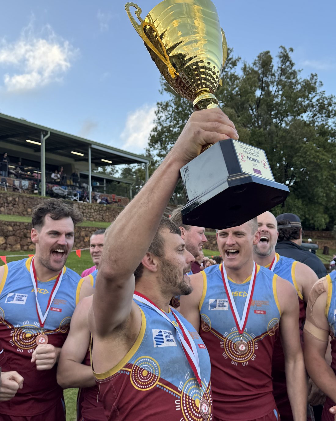 A Swan View player holding the premiership cup high as teammates in medals celebrate around him